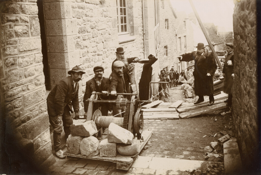 Ouvriers à l'œuvre dans la Grande Rue à hauteur du logis Saint-Aubert. Photographie, 1908 (Collection particulière). Après la séparation des Eglises et de l’Etat (1905), l’évêque diocésain confie l’organisation des pèlerinages et la célébration du XIIe centenaire de la fondation de l’abbaye (octobre 1908-octobre 1909) à son vicaire général, Mgr Lepetit. Ce dernier engage des travaux considérables sur plusieurs maisons du village dont il est devenu propriétaire.